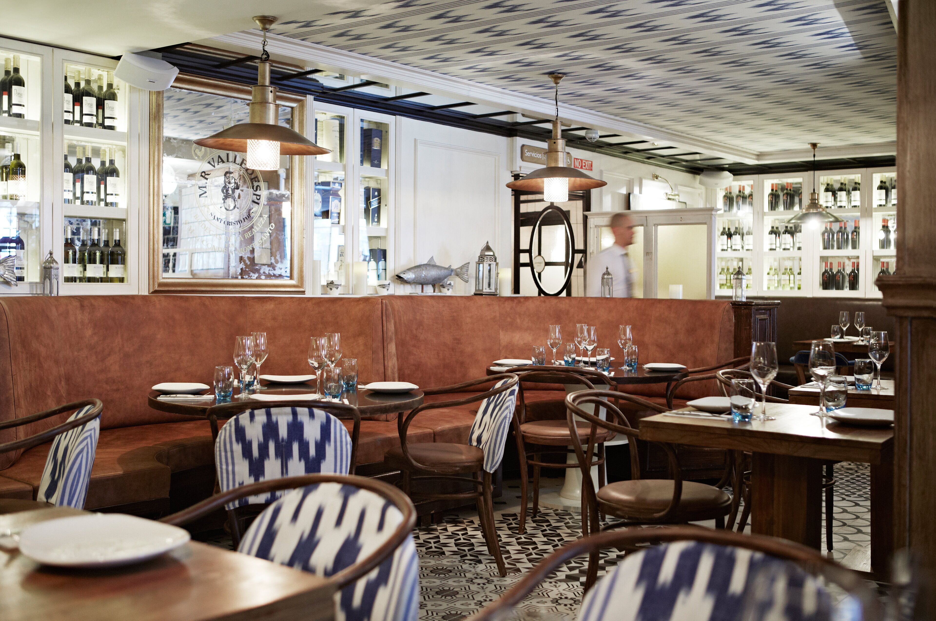 L’intérieur d’un restaurant avec des tables et des chaises en bois, un plafond orné de motifs et, au fond, un présentoir à vin.