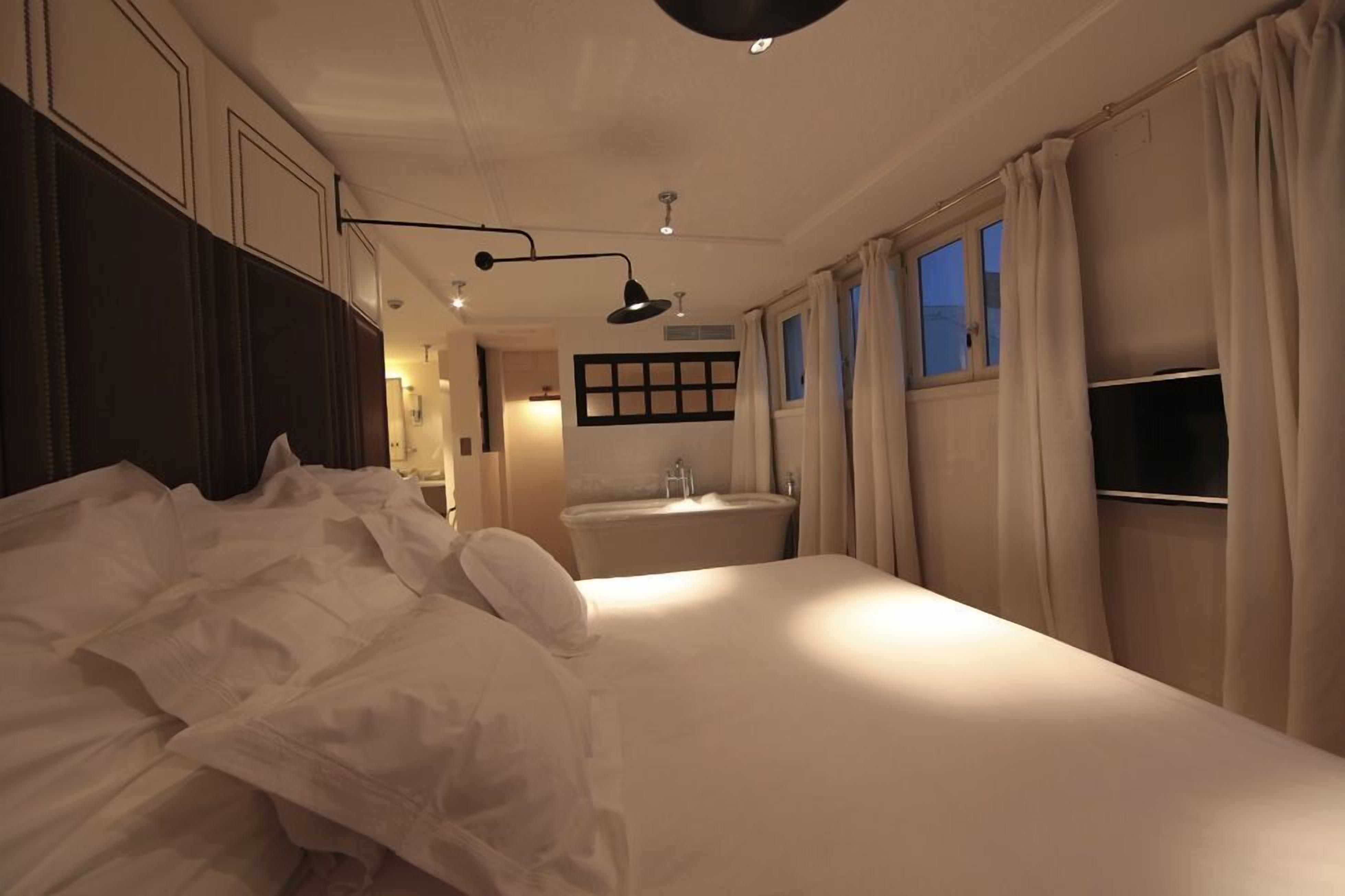 A hotel room with a large bed, a bedside table, a window with curtains, a wall-mounted TV, and a bathroom with a sink and mirror.