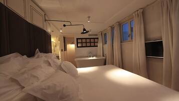 Una habitación de hotel con una cama grande, una mesita de noche, una ventana con cortinas, un televisor montado en la pared y un baño con lavamanos y espejo.
