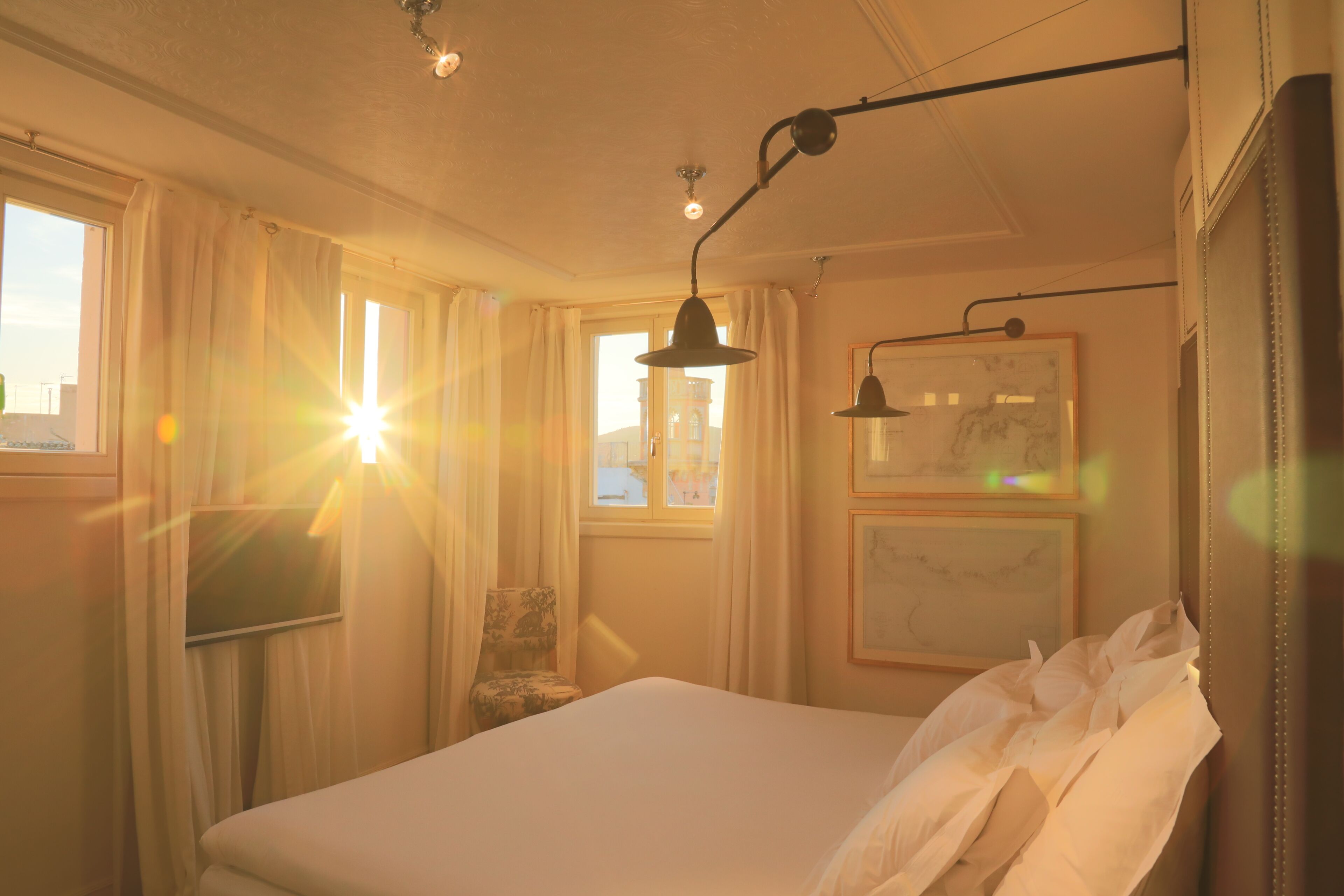 A hotel room with a bed, a chair, a desk, and a window with sunlight streaming in.