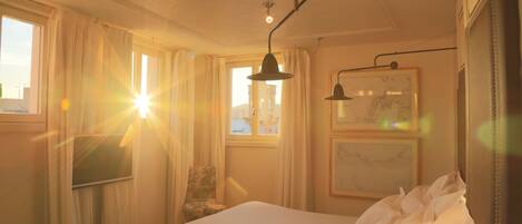 A hotel room with a bed, a chair, a desk, and a window with sunlight streaming in.