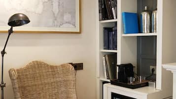 Un sillón de mimbre con un cojín estampado, una estantería con libros y objetos decorativos, un cuadro abstracto enmarcado y un refrigerador pequeño.