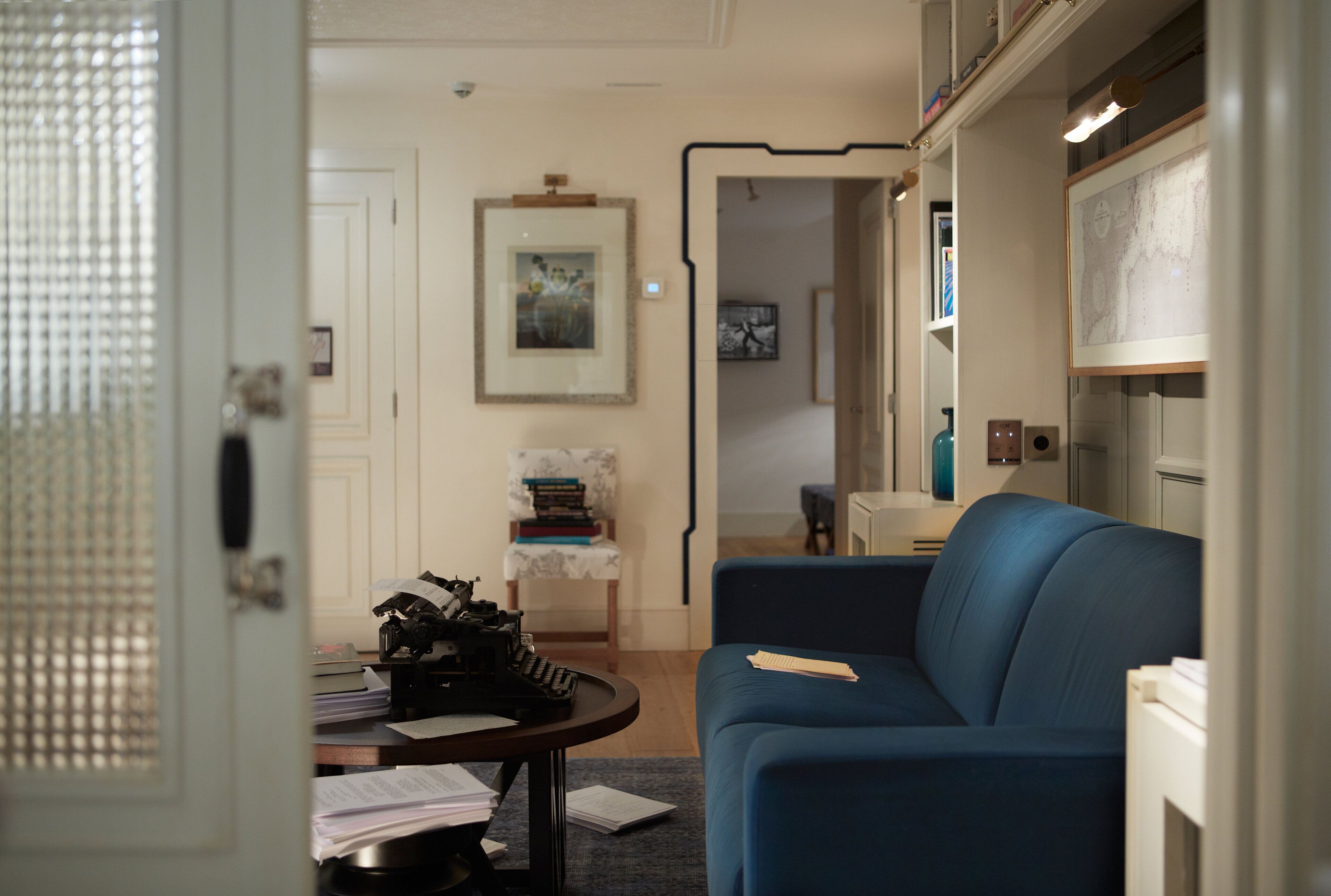 Una habitación con un sofá azul, una mesita con papeles, un cuadro en la pared y una puerta que da a otra habitación.