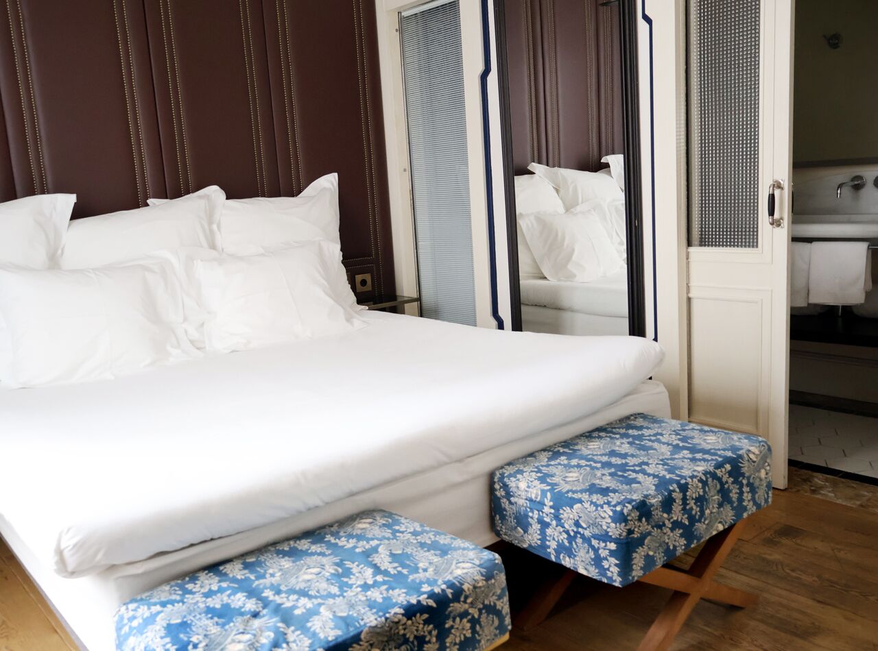 Uma cama bem arrumada com lençóis brancos, cabeceira de madeira e um par de pufes florais azuis.