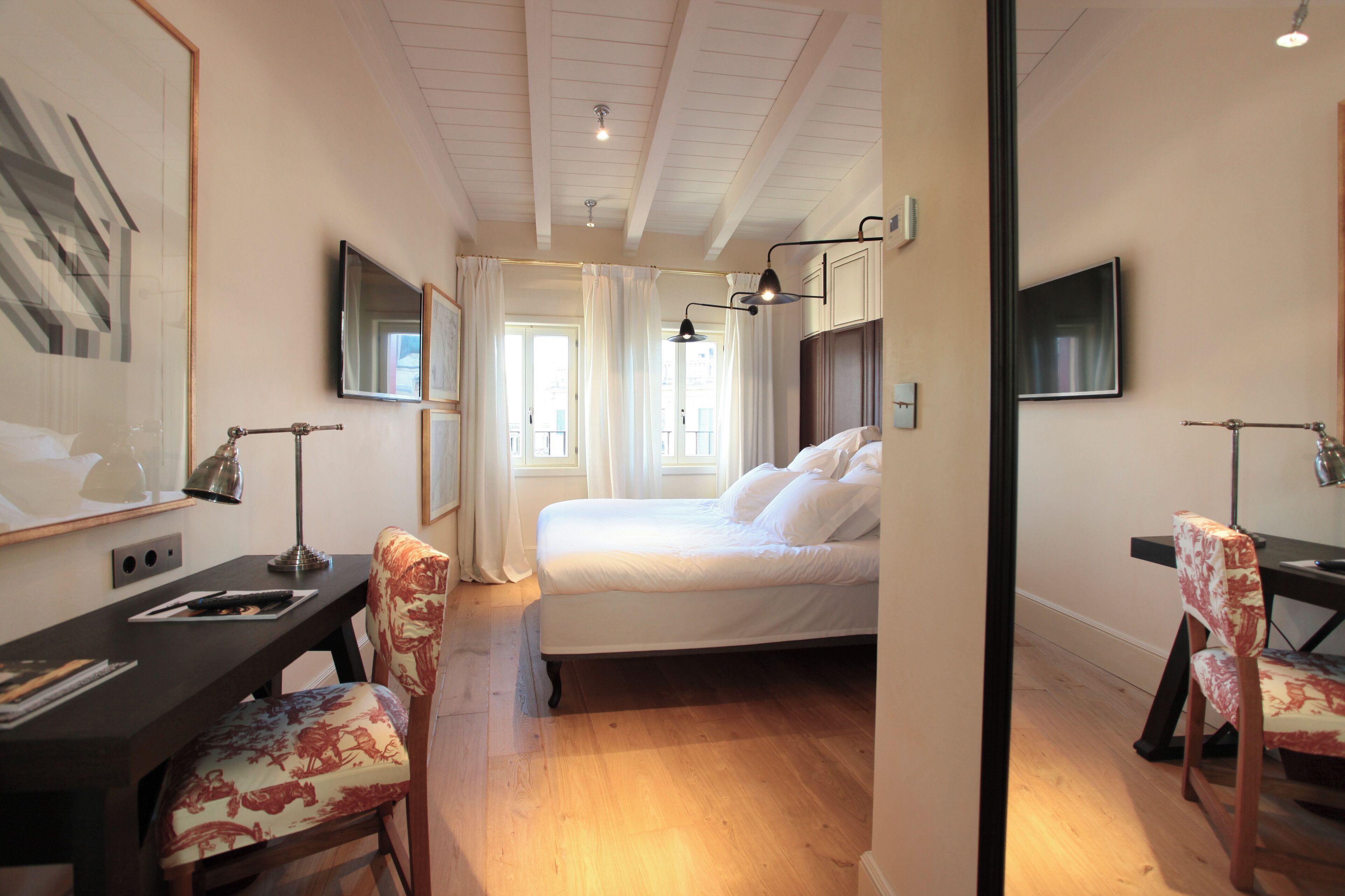 A hotel room with a bed, a desk, two chairs, a TV, and a window with curtains.