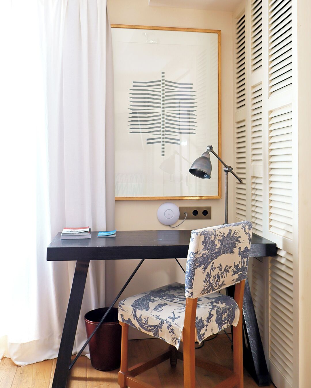 Een werkplek in de hoek met een bureau, stoel, lamp en een lijst met abstracte kunst.