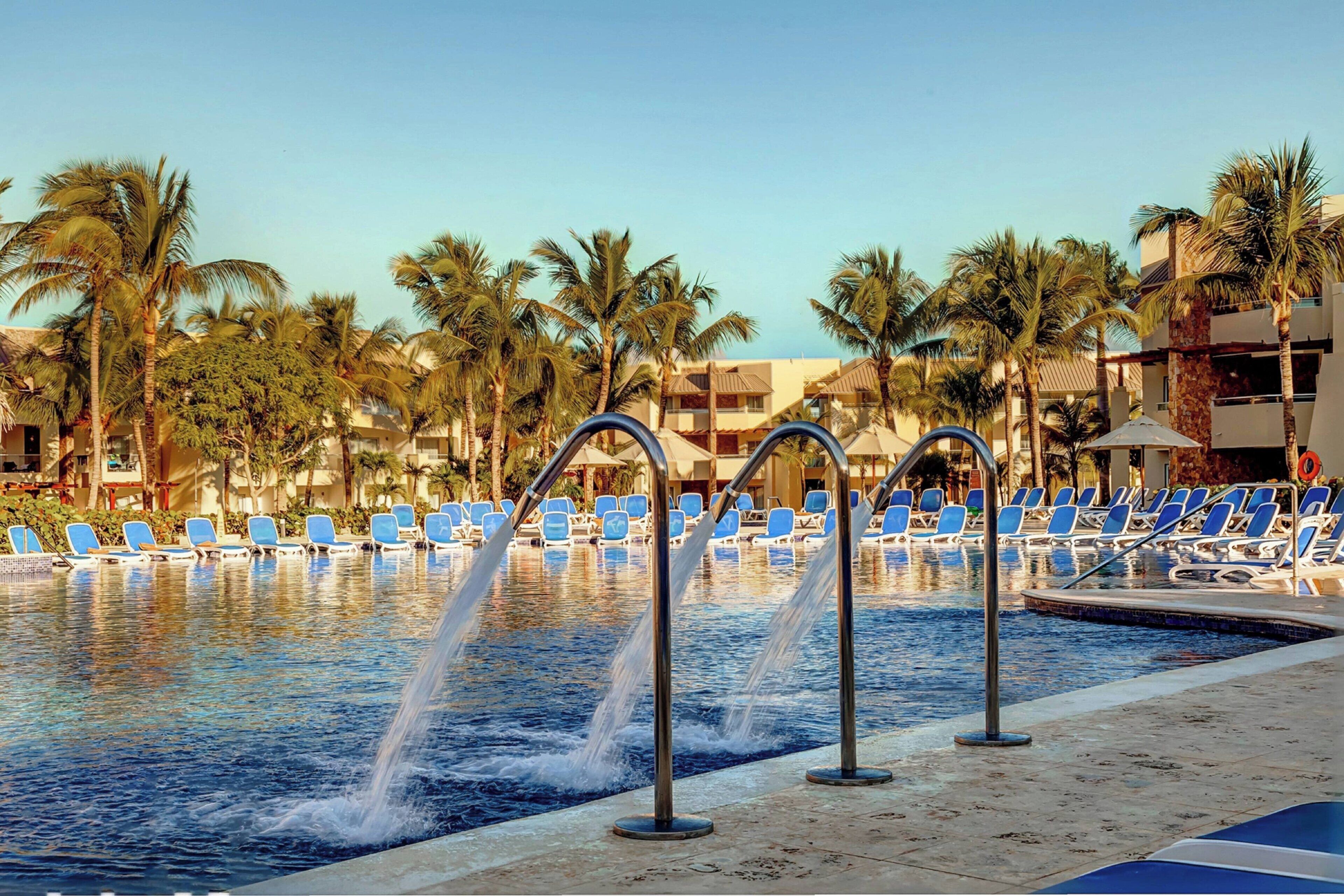 Zona de piscina con sillas azules y palmeras, rodeada de edificios.