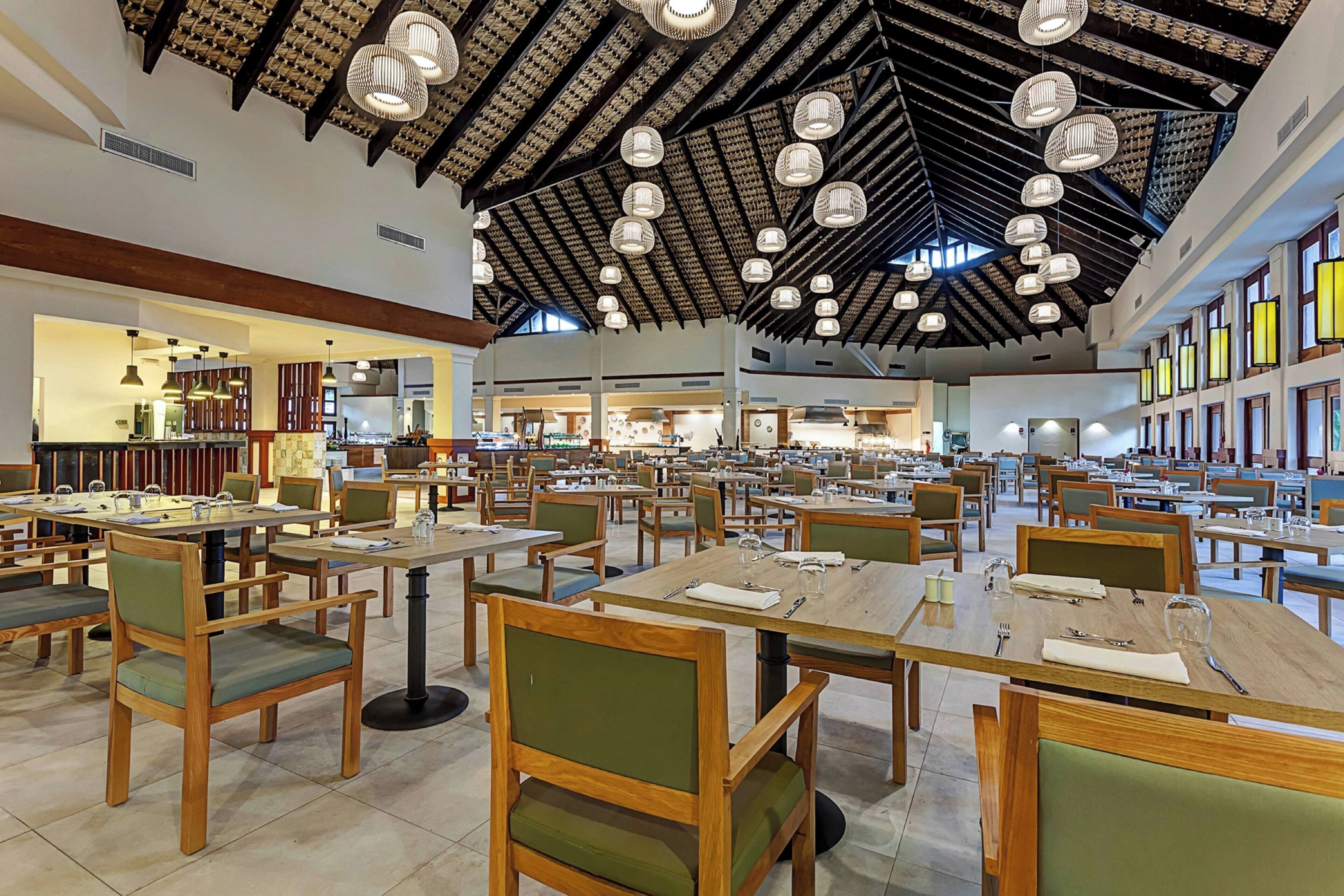A spacious dining area with multiple tables and chairs, a high ceiling with hanging lights, and a view of the kitchen area in the background.