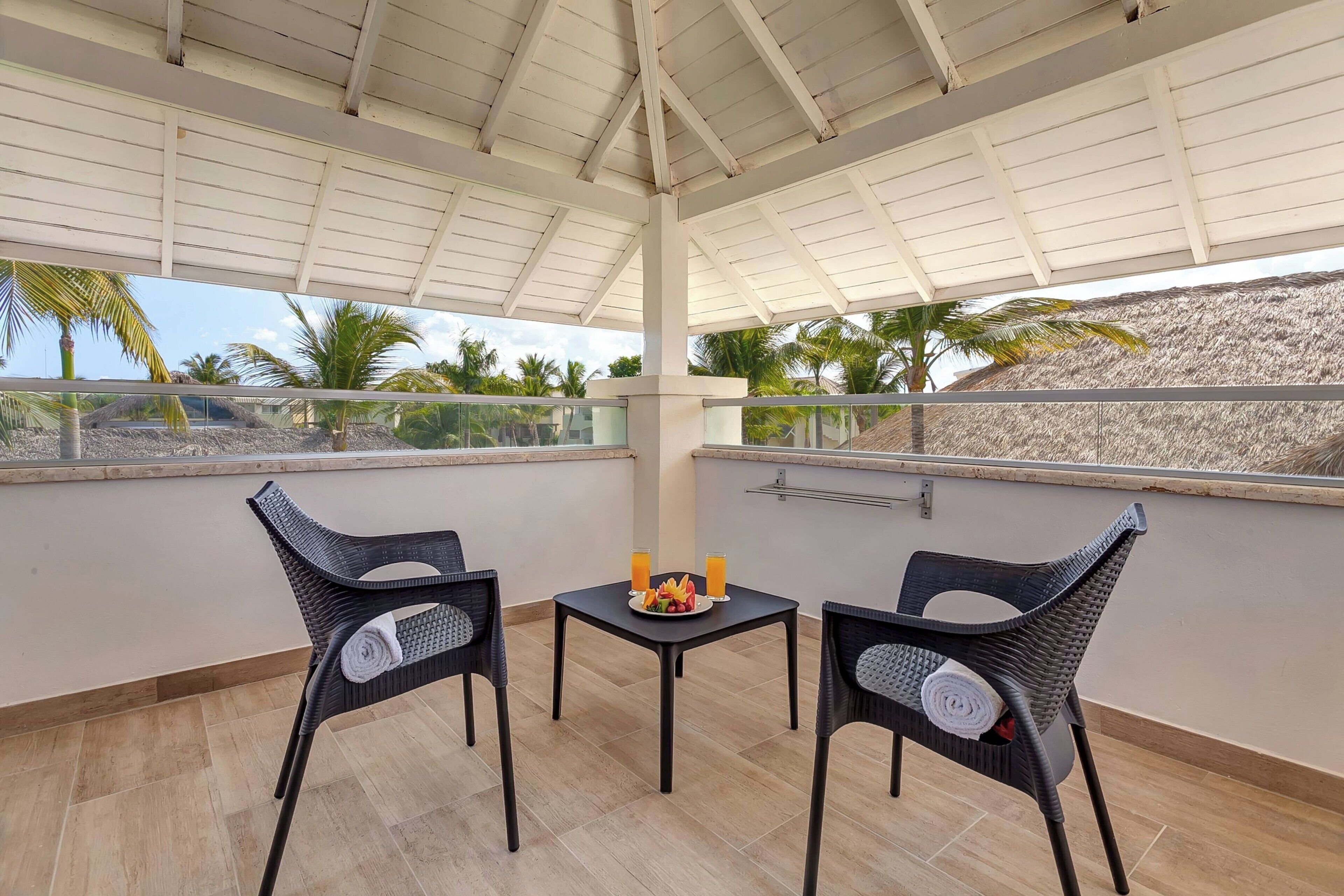 A covered balcony with two wicker chairs, a small table with a fruit plate and drinks, and a view of palm trees and a beach.
