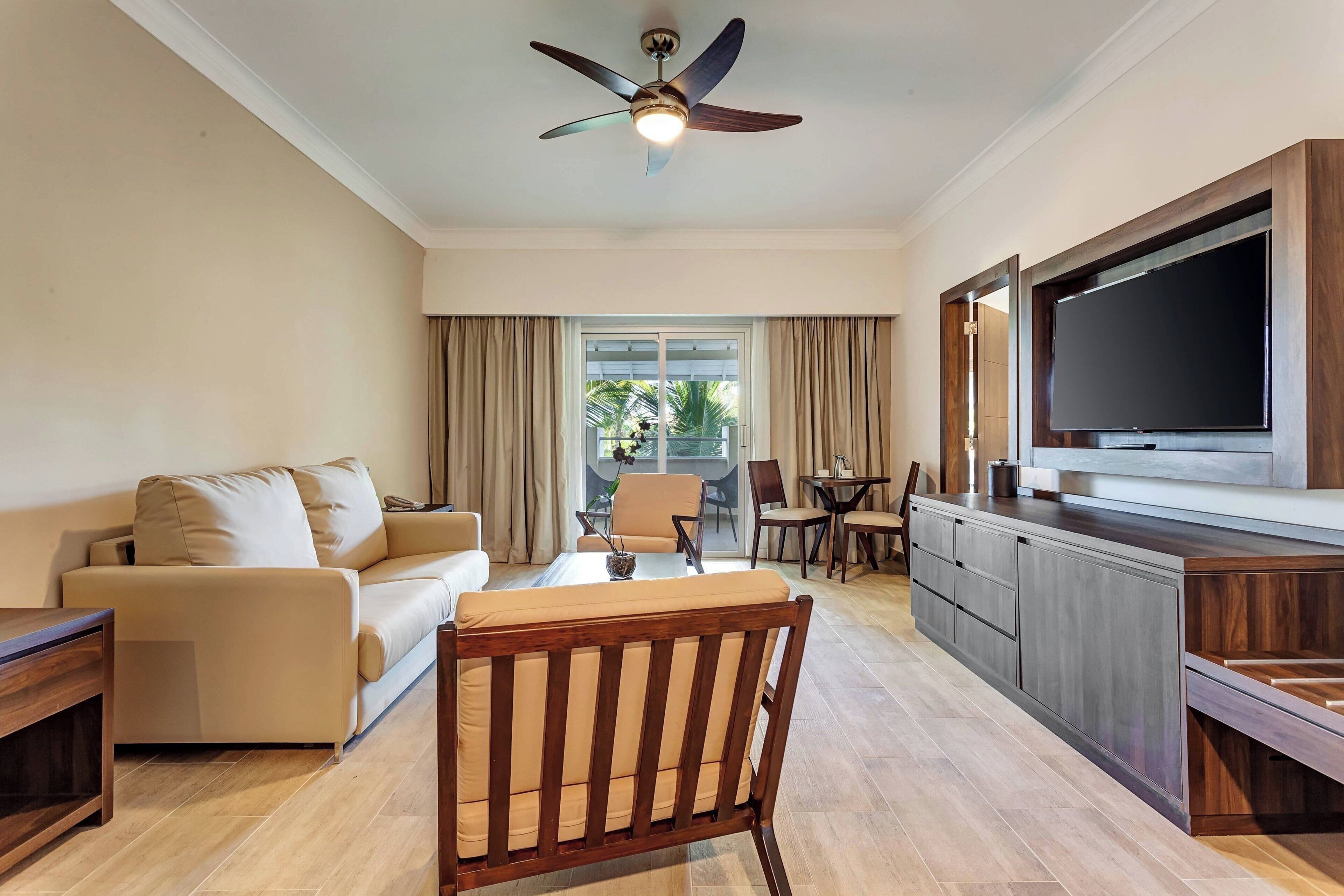 A modern living room with a sofa, a wooden chair, a dining table, a TV, and a ceiling fan.