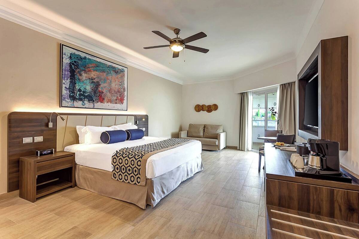 A hotel room with a large bed, a desk with a coffee maker, a ceiling fan, and a large window.
