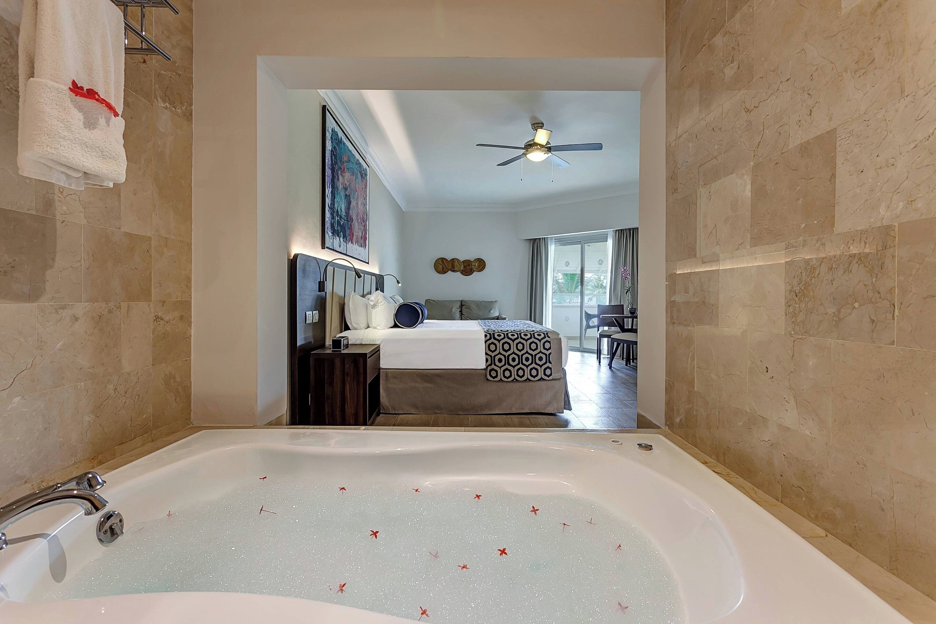 A modern bathroom with a large bathtub, a ceiling fan, and a bed in the background.
