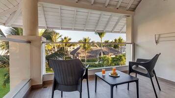 Un balcón con dos sillas y una mesa, con vistas a un resort tropical con palmeras y edificios de techo de paja.