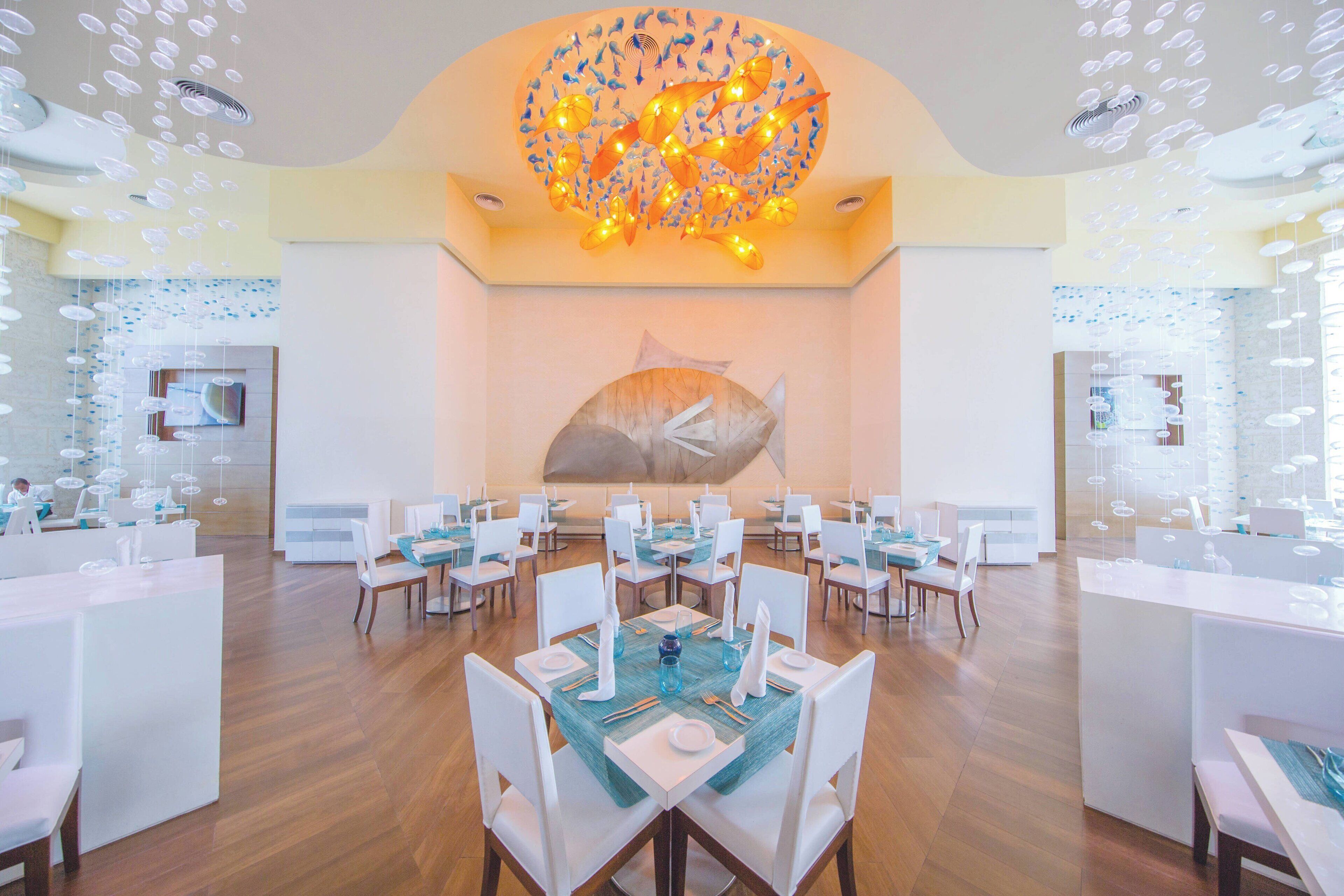 Un vaste espace dédié à la salle à manger, avec de nombreuses tables et chaises, une grande fresque représentant des poissons sur le mur et un luminaire au plafond au design original.