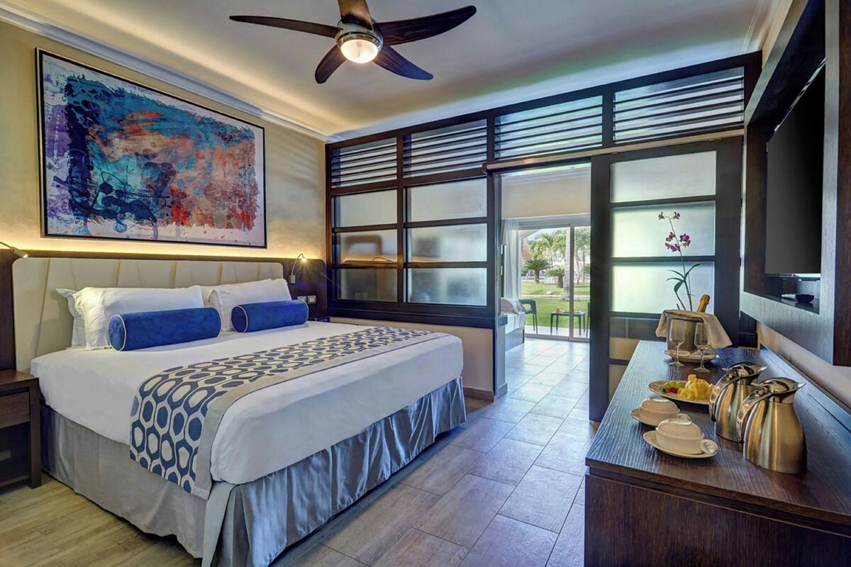 A modern hotel room with a large bed, a ceiling fan, a large window with blinds, and a dining area with breakfast items.