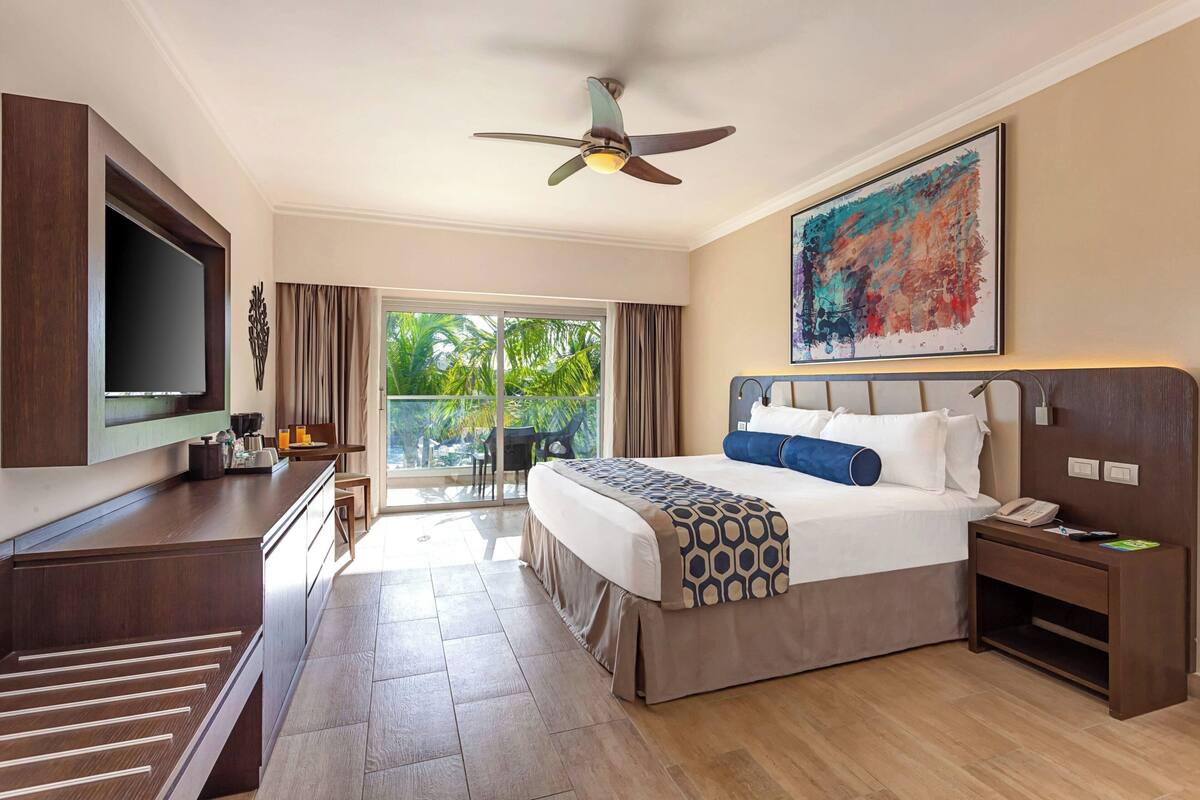 A hotel room with a bed, a desk, a television, a ceiling fan, and a view of the outdoors.
