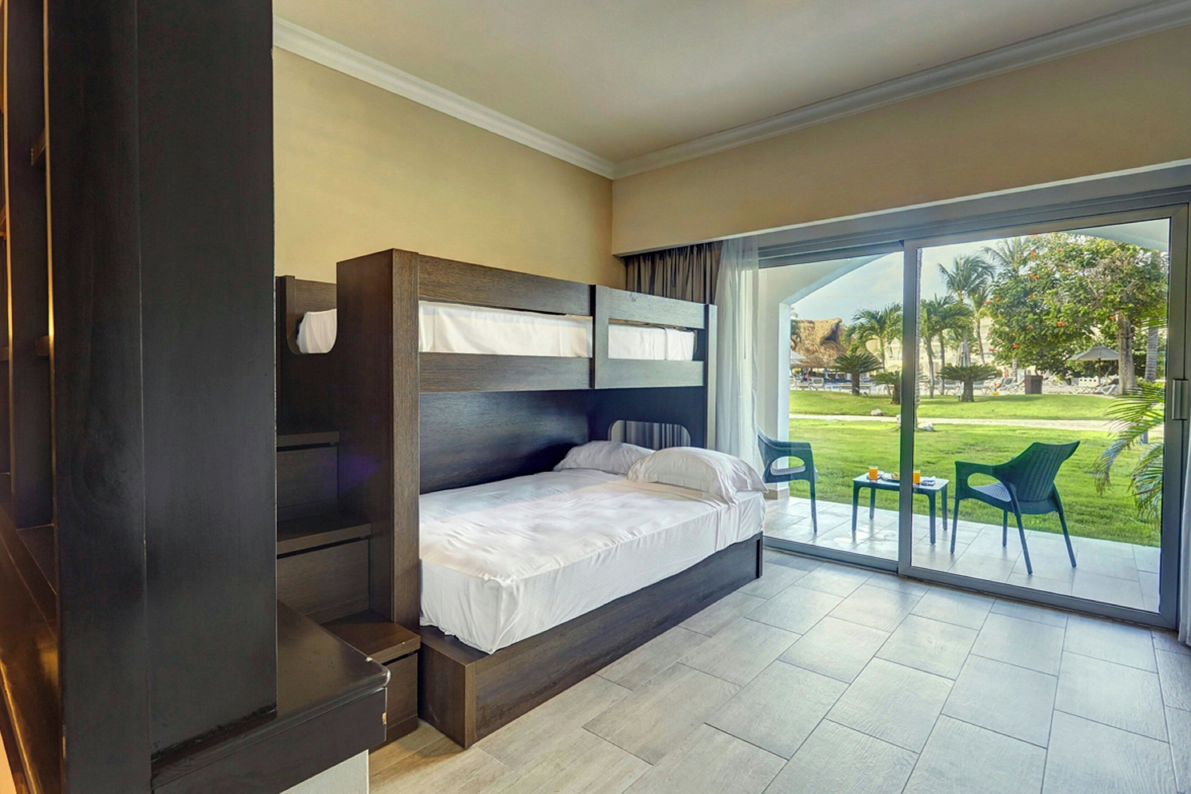 A hotel room with a bunk bed, a small desk, and a sliding glass door leading to a balcony with outdoor seating.