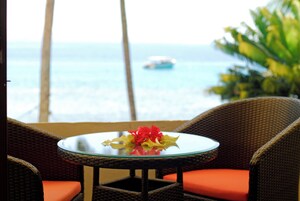 Una silla de mimbre con un cojín naranja, una mesita de vidrio con un arreglo floral y vista al océano con un barco a lo lejos.