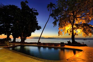 A pool area with lounge chairs, a palm tree, and a view of the sea at dusk. - Garden Island Resort (Taveuni Island East)