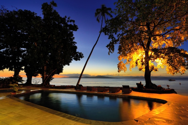 A pool area with lounge chairs, a palm tree, and a view of the sea at dusk. - Garden Island Resort (Taveuni Island East)