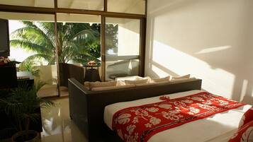 A modern hotel room with a large bed, a wicker sofa, a small table, and a view of palm trees through large sliding glass doors.