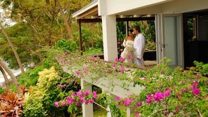 Un espacio al aire libre cubierto con una estructura blanca, abundante follaje y flores rosadas.