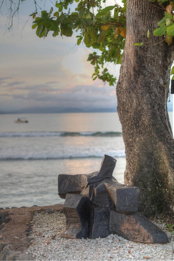 A wooden sculpture by a tree on a pebbled beach with a view of the ocean and a boat in the distance. - Garden Island Resort (Taveuni Island East)