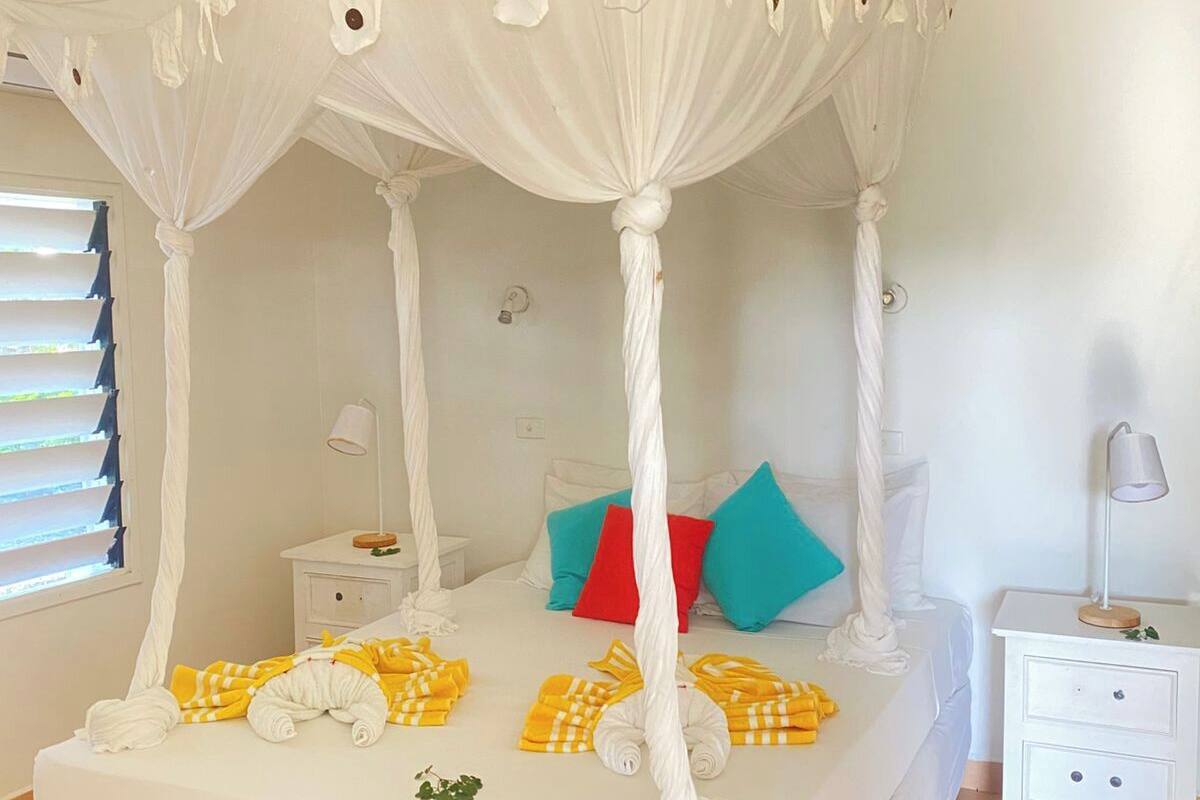 A four-poster canopy bed with white bedding and yellow towels, a bedside table with a lamp, and a window with blinds.