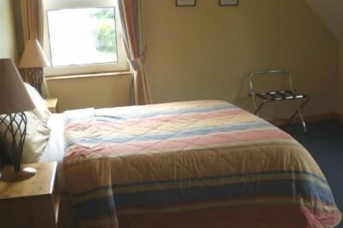 A hotel room with a bed, a nightstand, a lamp, a window with curtains, and a chair.