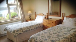 A bedroom with two beds, a nightstand, a dresser, a lamp, and a window with a view of greenery.