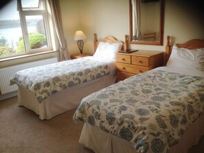 A bedroom with two beds, a nightstand, a dresser, a lamp, and a window with a view of greenery.