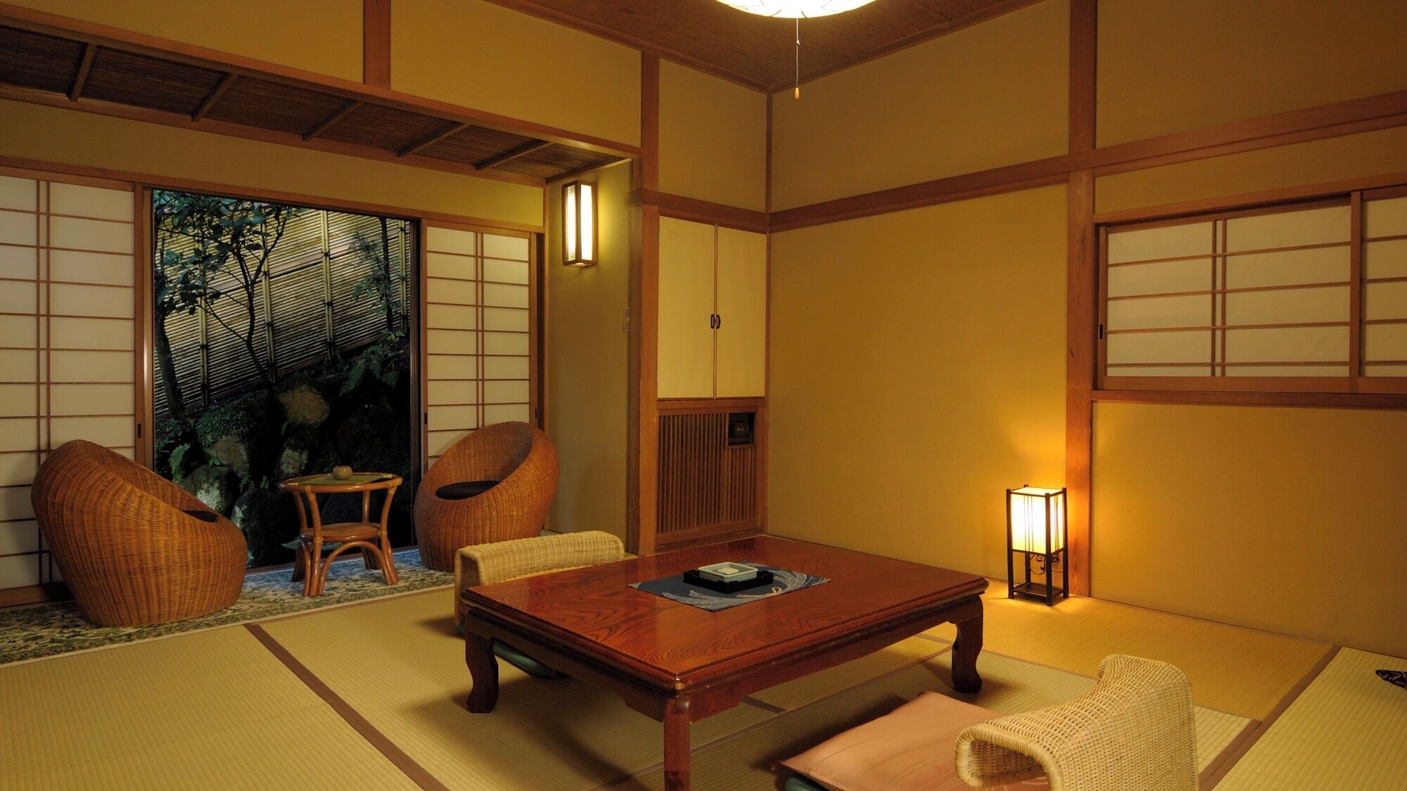 A traditional Japanese room with tatami flooring, a low wooden table, two wicker chairs, a small table with a tea set, and sliding doors with shoji screens.
