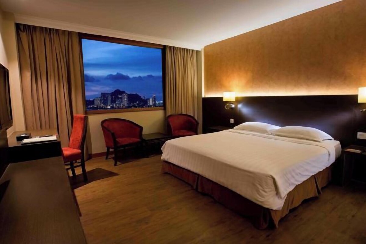 A hotel room with a bed, a desk, a chair, and a view of a cityscape at dusk.