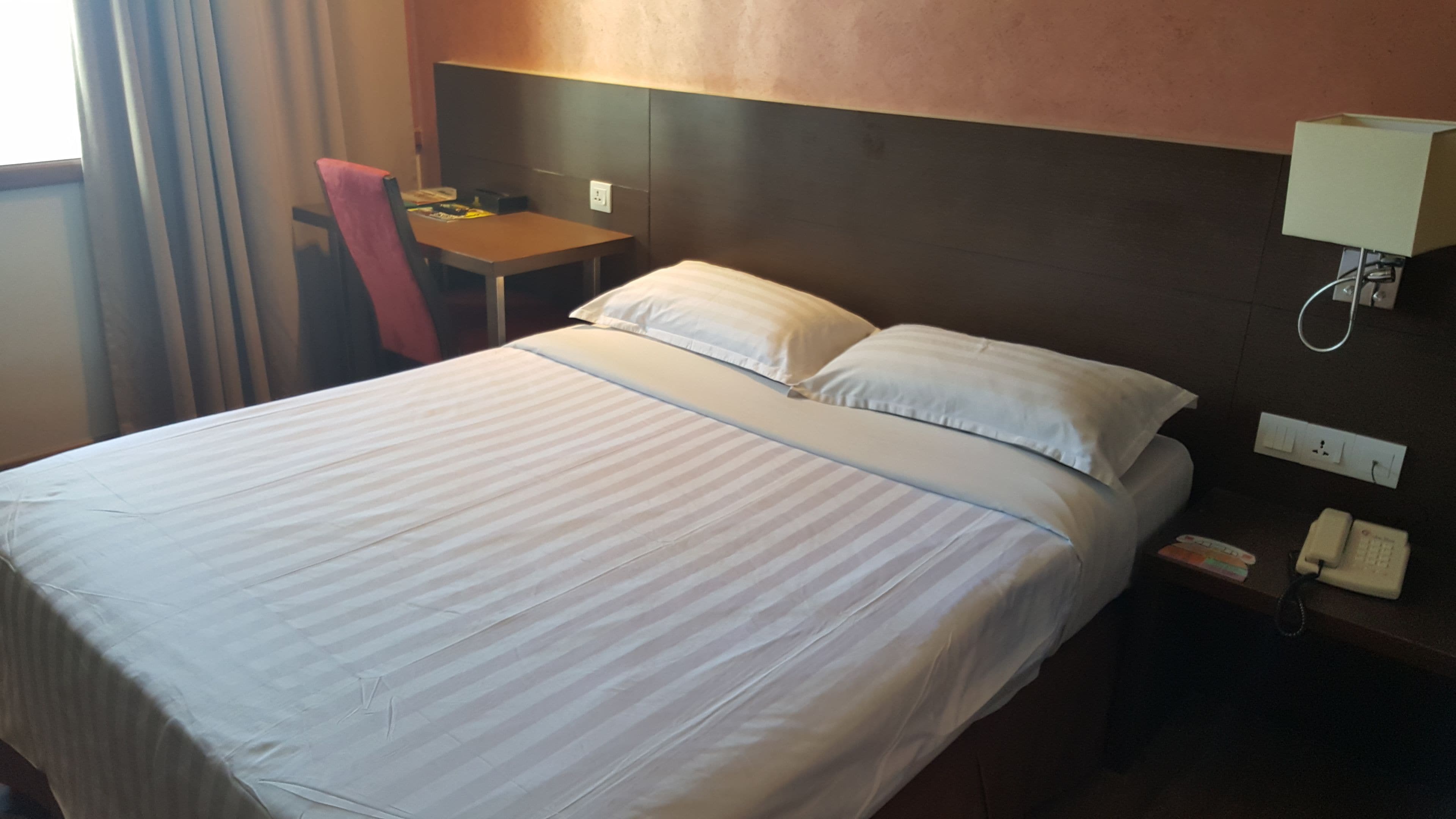 A hotel room with a bed, a desk, a chair, a lamp, and a phone on the nightstand.