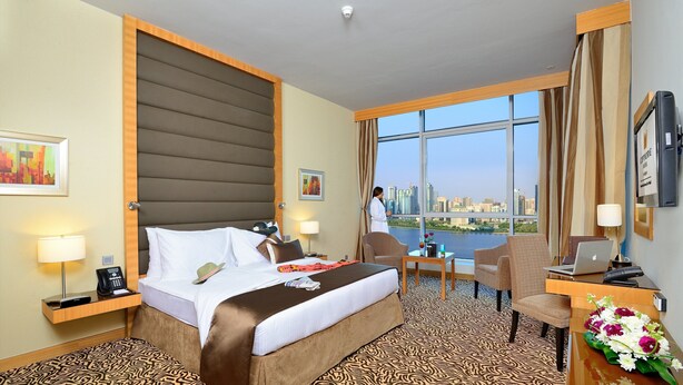 A hotel room with a large bed, a desk with a laptop, a chair, and a view of a cityscape. - Copthorne Hotel Sharjah (Sharjah)