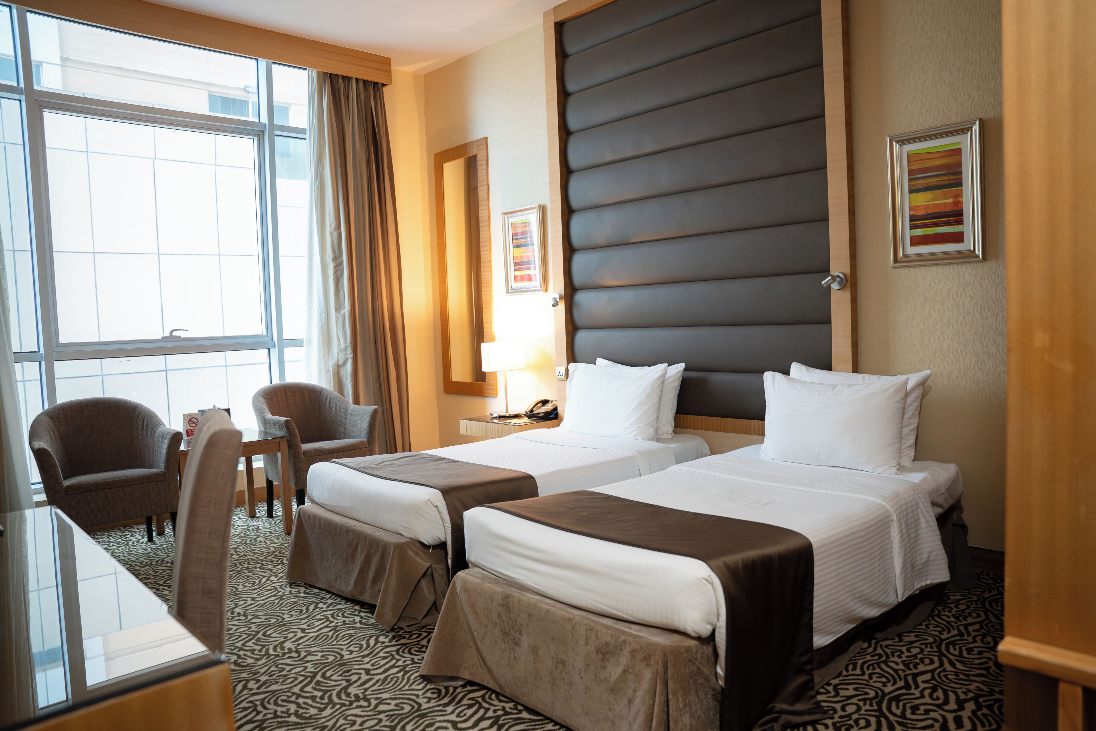 Habitación de hotel con dos camas, un escritorio, sillas, un ventanal grande y alfombra estampada.
