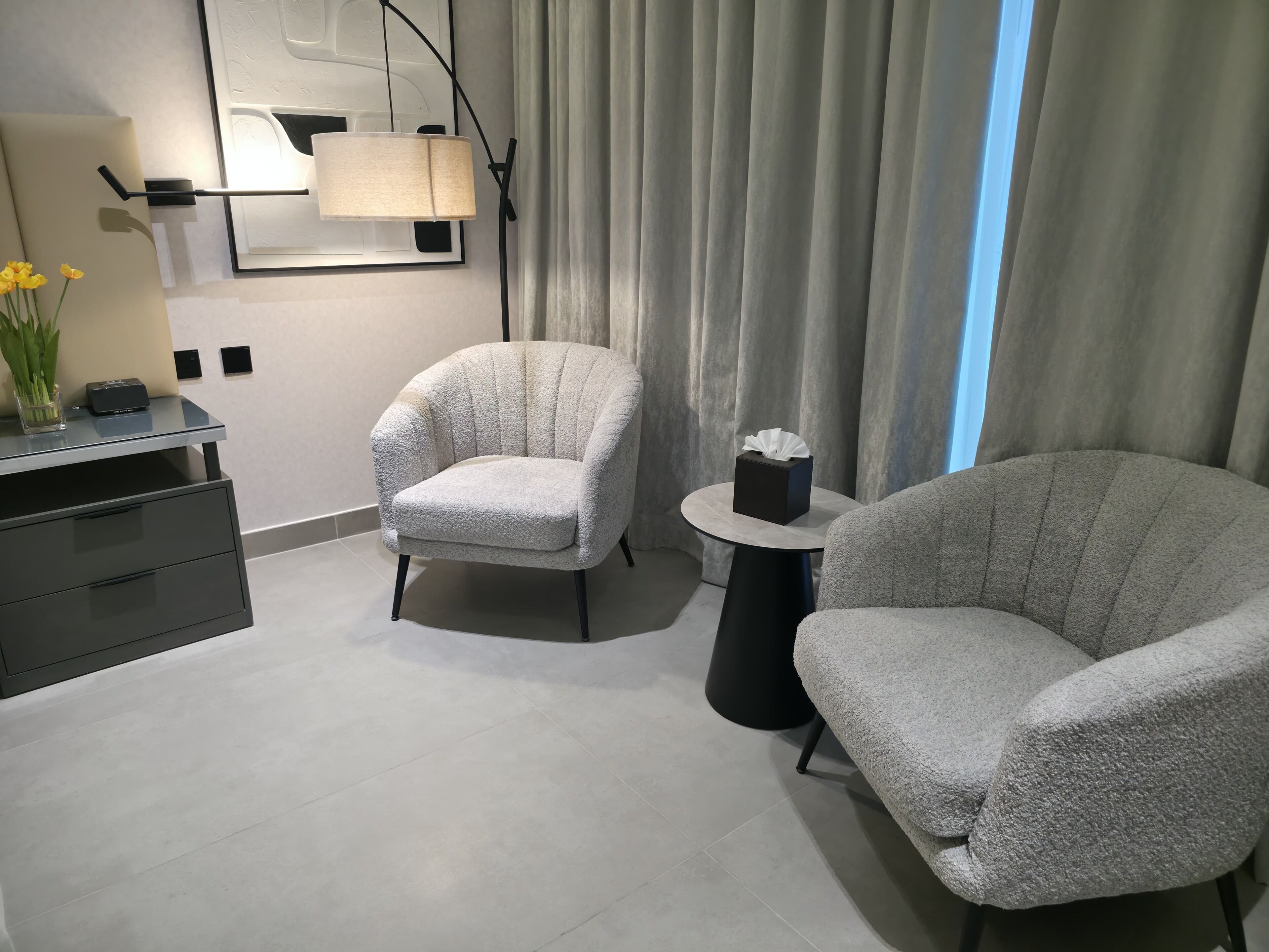Ein modernes Zimmer mit zwei Sesseln, einem kleinen Tisch mit Taschentuchbox, einer Lampe und einem Schreibtisch mit Blumenvase.