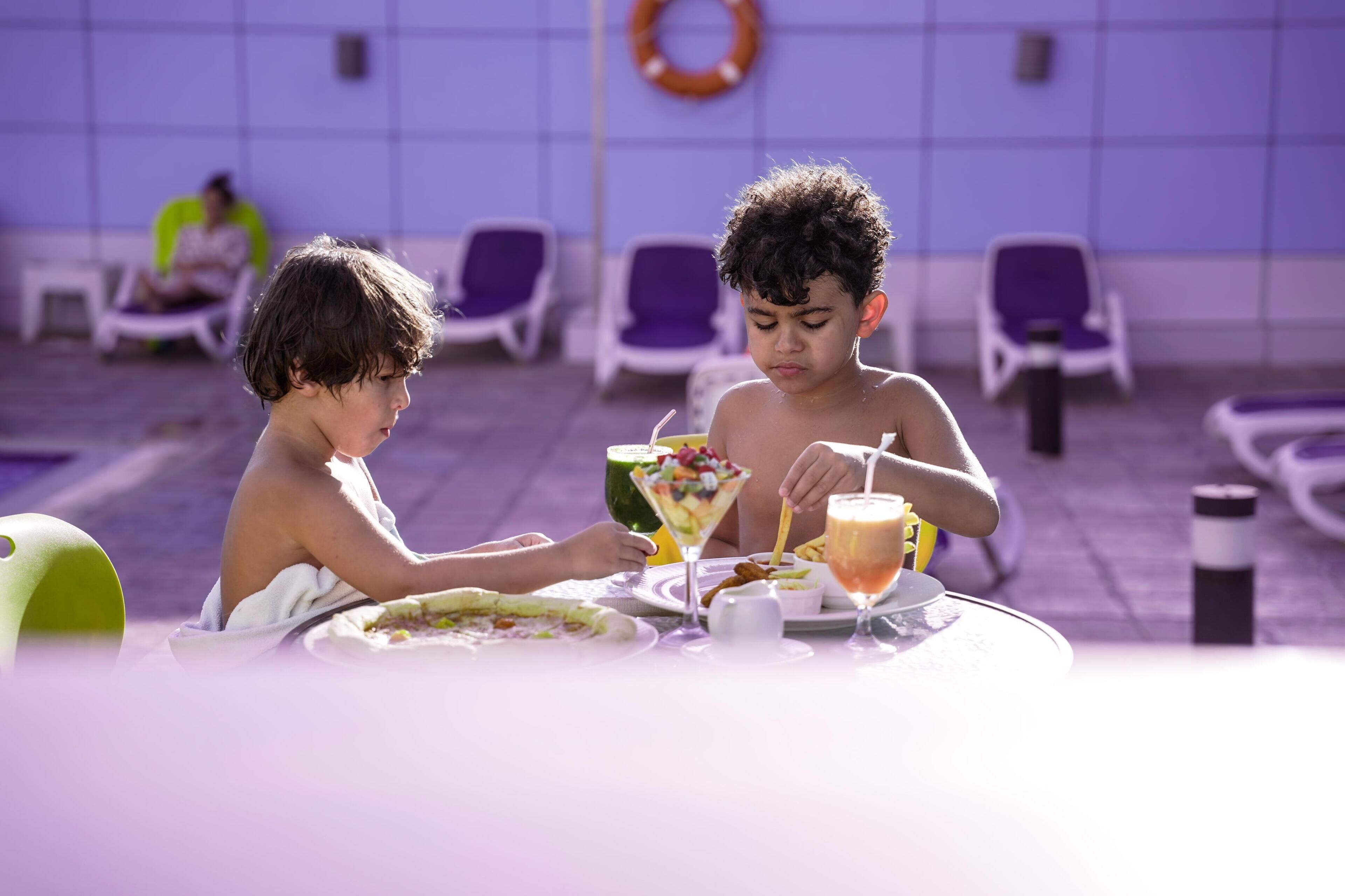 Deux enfants sont assis à une table et déjeunent, avec une piscine en arrière-plan.
