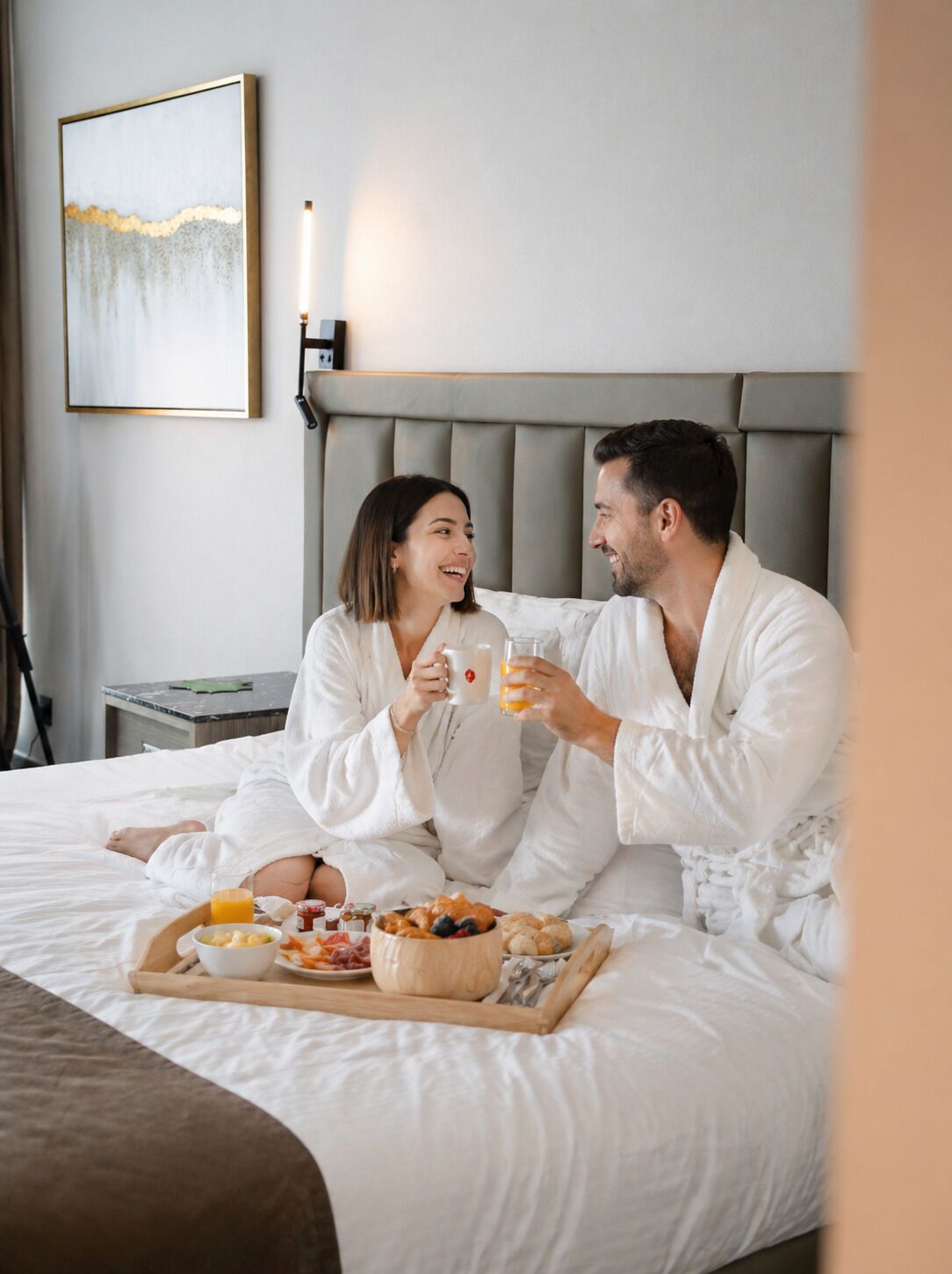 Ein Paar in weißen Gewändern sitzt auf einem Bett mit Frühstücksgebäck, einem Nachttisch und einem Gemälde an der Wand.