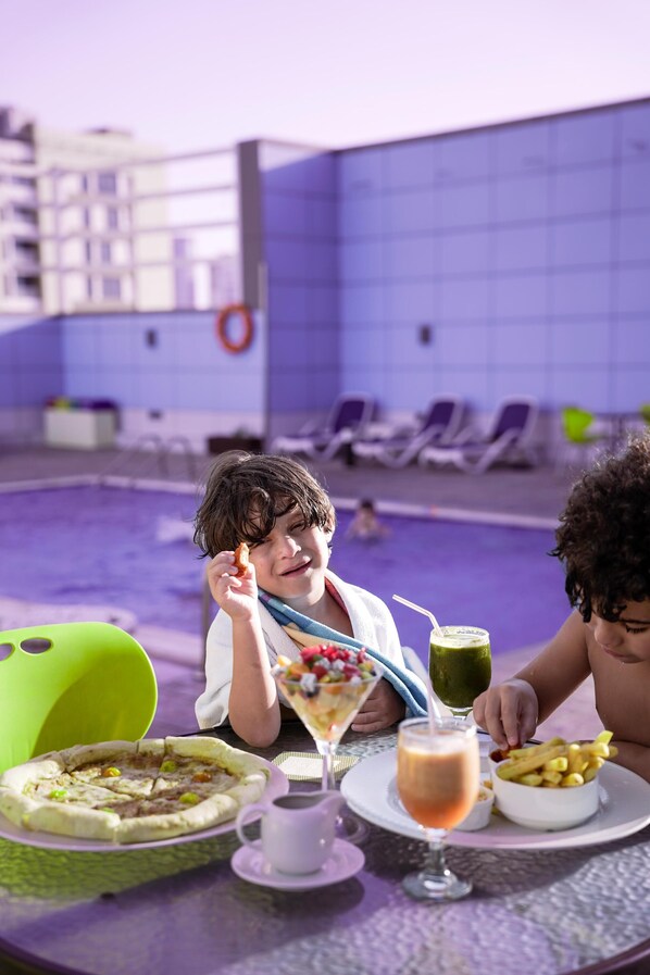 Zona de comedor junto a la piscina con una mesa para dos, mostrando una pizza, una bebida verde y un postre.