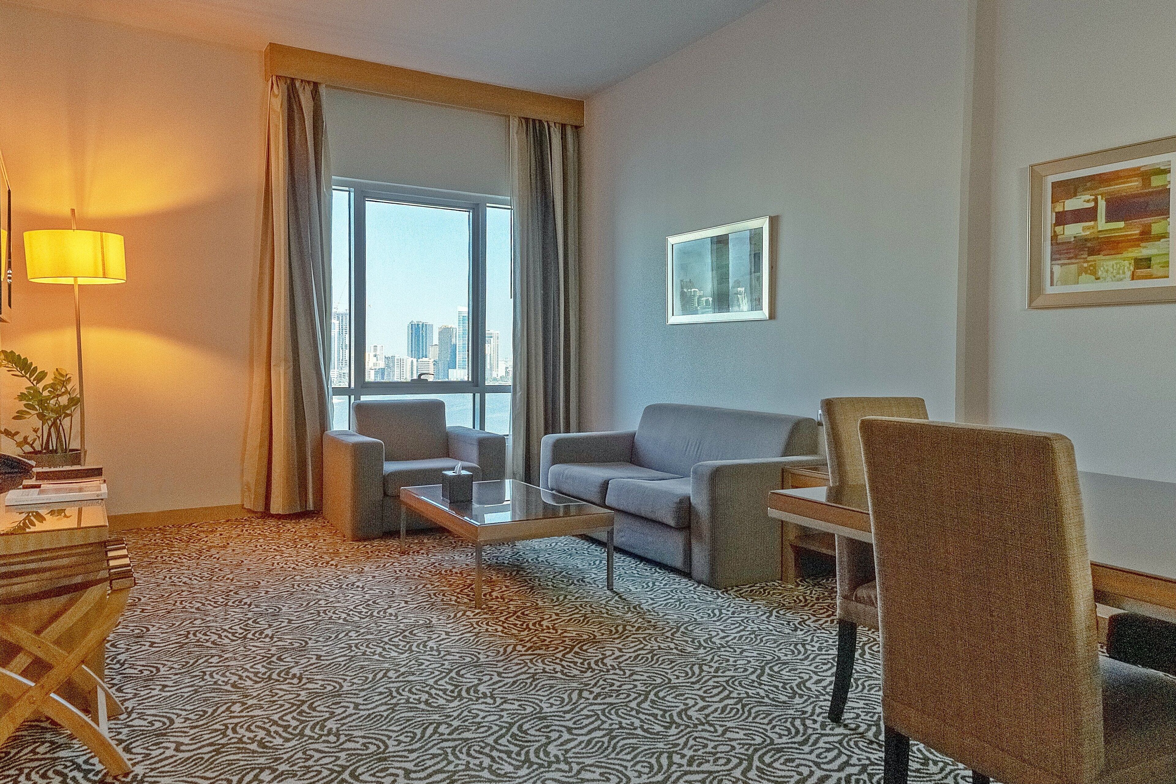 Ein Hotelzimmer mit Teppichmuster, einer Couch, einem Sessel, einem Couchtisch, einer Lampe, einem Fenster mit Vorhängen und einem gerahmten Bild an der Wand.