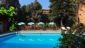A swimming pool with lounge chairs and umbrellas, surrounded by trees and a brick building. - Hotel Greenwich Village (Lalitpur)
