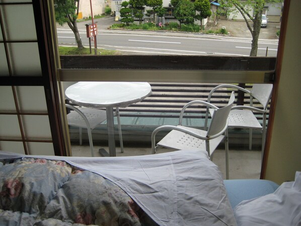 A room with a bed, a small table, and two chairs. There is a window with a view of a street and buildings.
