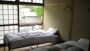 A room with two beds, one with a floral bedspread and another with a plain white bedspread. There is a wooden chair and a wooden bed frame. A window with a view of trees and buildings is visible. - Towadako Backpackers (Towada)