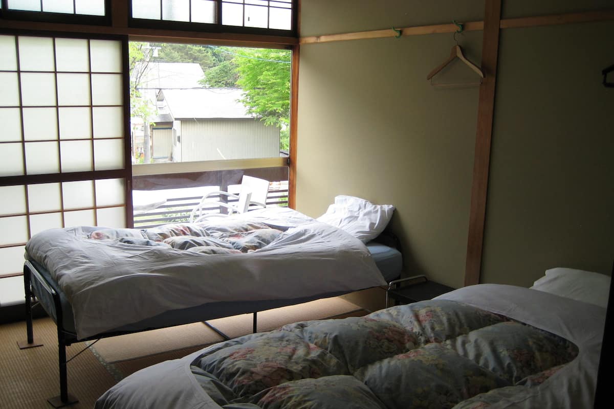 Una habitación con dos camas, una con cubrecama floral y otra con cubrecama blanca lisa. Hay una silla de madera y un cabecero de madera. Se ve una ventana con vistas a árboles y edificios.