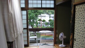 A room with tatami flooring, a futon bed, a small table with chairs, a standing fan, and a view of the outside through a shoji screen. - Towadako Backpackers (Towada)