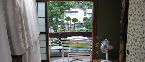 A room with tatami flooring, a futon bed, a small table with chairs, a standing fan, and a view of the outside through a shoji screen.