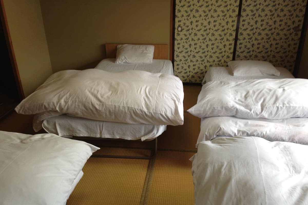 Una habitación con varias camas, cada una con ropa de cama blanca y cabecero de madera. El suelo es de tatami y la pared tiene un estampado floral.