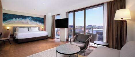 A hotel room with a large bed, a flat-screen TV, a sofa, a small table, and a view of the city.