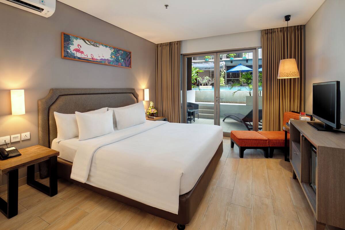 A hotel room with a bed, bedside table, TV, and a view of the outdoor pool area.